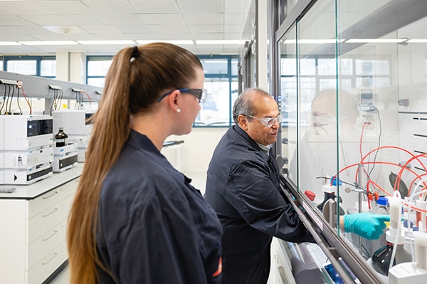 colleague watches a lab worker perform experiment under a hood